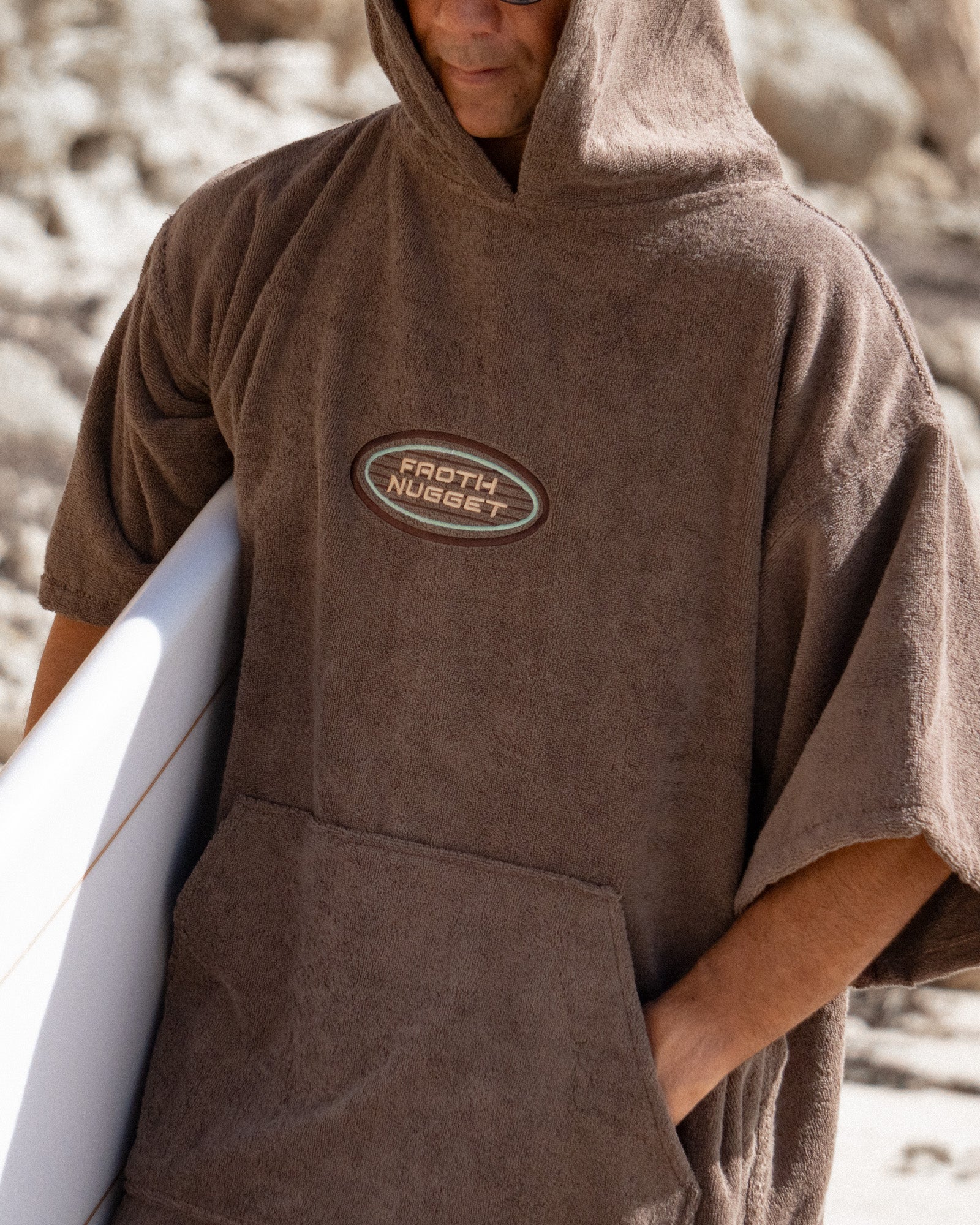 A man on a beach wearing a brown hooded towel poncho with a front pocket and a visible Froth Nugget logo patch on the chest, holding a white surfboard.